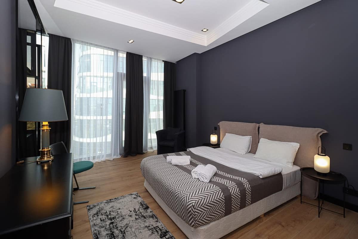 The bedroom includes a king-size bed, a black nightstand, bedside lamps, and various seating options.